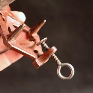 May include: A rusty brown metal clamp with a screw and a ring. The clamp has two pointed prongs and a screw mechanism for tightening. The background is dark.