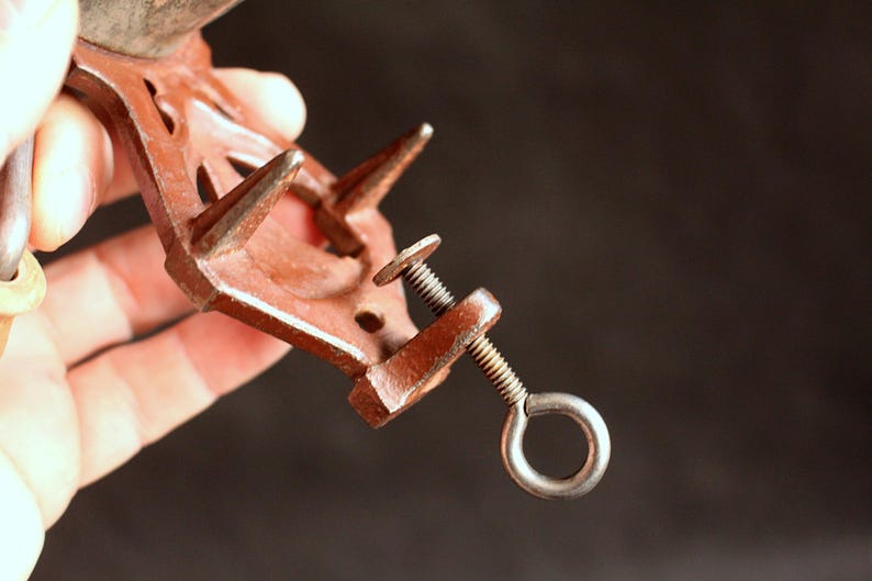 May include: A rusty brown metal clamp with a screw and a ring. The clamp has two pointed prongs and a screw mechanism for tightening. The background is dark.
