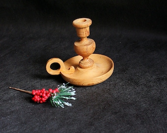 May include: A handcrafted wooden candlestick holder with a round base and a handle. The candlestick is made of light-colored wood and features a decorative design. A sprig of red berries and green foliage with white accents rests beside the candlestick.
