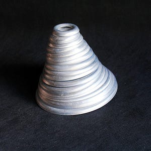 Puede incluir: Un recipiente plateado en forma de cono con tapa. El recipiente est&aacute; hecho de anillos circulares apilados, creando una superficie texturizada y acanalada. El objeto se encuentra sobre una superficie oscura y texturizada.