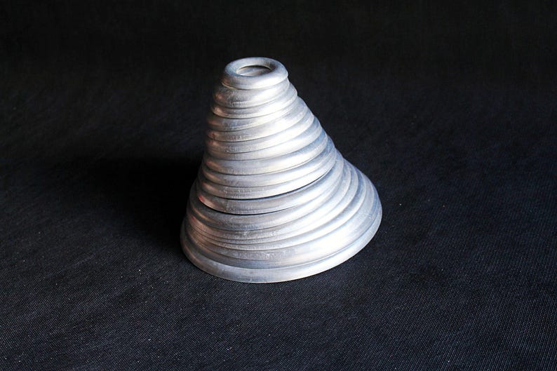 Puede incluir: Un recipiente plateado en forma de cono con tapa. El recipiente est&aacute; hecho de anillos circulares apilados, creando una superficie texturizada y acanalada. El objeto se encuentra sobre una superficie oscura y texturizada.