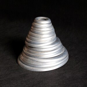 Puede incluir: Una escultura plateada en forma de cono compuesta por anillos met&aacute;licos circulares apilados. Los anillos disminuyen de tama&ntilde;o a medida que ascienden, creando una apariencia texturizada y en capas. La escultura est&aacute; sobre un fondo oscuro.
