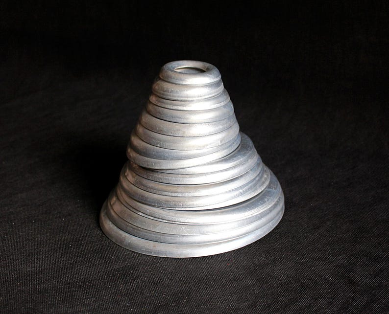Puede incluir: Una escultura plateada en forma de cono compuesta por anillos met&aacute;licos circulares apilados. Los anillos disminuyen de tama&ntilde;o a medida que ascienden, creando una apariencia texturizada y en capas. La escultura est&aacute; sobre un fondo oscuro.