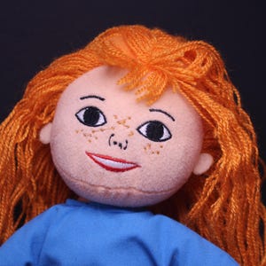 Pode incluir: Boneca feita &agrave; m&atilde;o com tom de pele claro, sardas e cabelo de fio laranja. A boneca tem olhos pretos bordados, um sorriso vermelho e est&aacute; vestida com uma blusa azul. O fundo &eacute; preto liso.