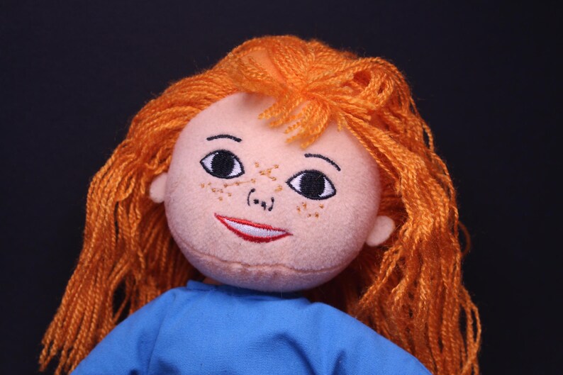 Pode incluir: Boneca feita &agrave; m&atilde;o com tom de pele claro, sardas e cabelo de fio laranja. A boneca tem olhos pretos bordados, um sorriso vermelho e est&aacute; vestida com uma blusa azul. O fundo &eacute; preto liso.
