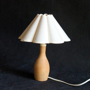 May include: A small, vintage table lamp with a cream-colored, pleated lampshade. The base is a light brown, wooden bottle shape. A white cord extends from the base against a black background.