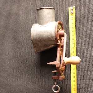 May include: An antique metal food grinder with a crank handle and clamp. The grinder is silver with a rusty brown handle and clamp. A yellow measuring tape is next to the grinder.