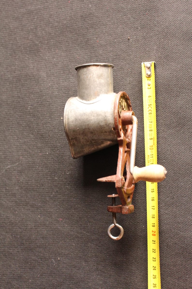 May include: An antique metal food grinder with a crank handle and clamp. The grinder is silver with a rusty brown handle and clamp. A yellow measuring tape is next to the grinder.