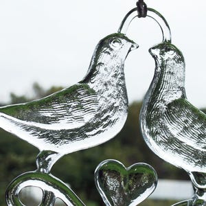 Puede incluir: Dos palomas de vidrio transparente con un dise&ntilde;o en forma de coraz&oacute;n, bes&aacute;ndose. Las palomas est&aacute;n suspendidas de una cuerda negra.
