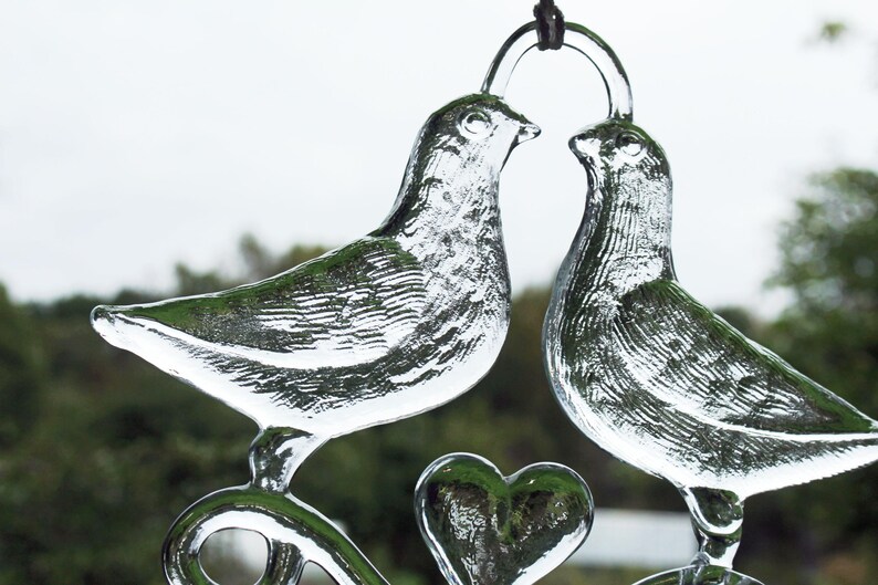 Puede incluir: Dos palomas de vidrio transparente con un dise&ntilde;o en forma de coraz&oacute;n, bes&aacute;ndose. Las palomas est&aacute;n suspendidas de una cuerda negra.