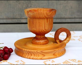 May include: A wooden candle holder with a round base and a cup-shaped top. The holder is made of light brown wood with a natural grain pattern.