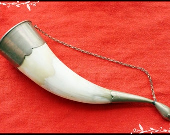 May include: A white horn drinking cup with silver accents and a chain. The horn is on a red fabric background.