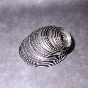 Puede incluir: Una pila de arandelas met&aacute;licas plateadas dispuestas en espiral. Las arandelas, de tama&ntilde;o uniforme, tienen un agujero central. La disposici&oacute;n crea una forma curva tridimensional sobre un fondo gris.