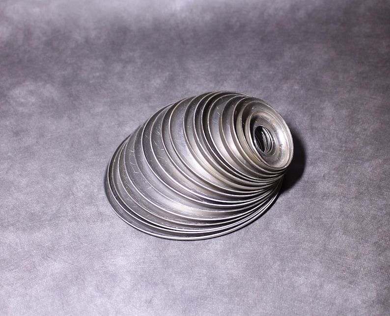 Puede incluir: Una pila de arandelas met&aacute;licas plateadas dispuestas en espiral. Las arandelas, de tama&ntilde;o uniforme, tienen un agujero central. La disposici&oacute;n crea una forma curva tridimensional sobre un fondo gris.
