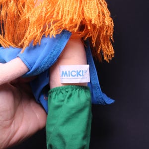 Pode incluir: Uma boneca vintage com cabelo de fio laranja, usando uma blusa azul e cal&ccedil;as verdes. A boneca tem uma etiqueta que diz "MICKI". Um brinquedo cl&aacute;ssico.