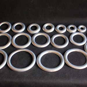 Puede incluir: Conjunto de anillos met&aacute;licos plateados de diferentes tama&ntilde;os, dispuestos sobre una superficie negra. Los anillos son circulares con un acabado liso y mate. Perfectos para manualidades y bisuter&iacute;a.
