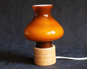 Puede incluir: Una pequeña lámpara de mesa vintage con una pantalla de cristal ámbar cálido y una base de madera. La pantalla tiene una forma redondeada, como de hongo, y se asienta sobre una base cilíndrica de madera. Un cable blanco se extiende desde la base.