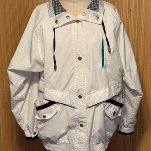 May include: White jacket with navy blue and teal accents. Features a plaid collar, gold buttons, and two front pockets. The jacket has a drawstring and a sporty, casual style.