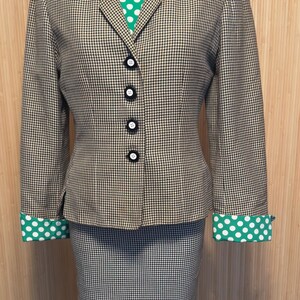 May include: A two-piece suit featuring a blazer and a skirt in a black and tan houndstooth pattern. The blazer has four black buttons and green cuffs with white polka dots. A green shirt with white polka dots is visible underneath the blazer collar.