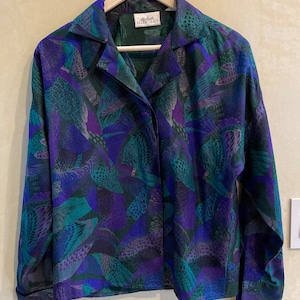 May include: A long-sleeved button-down shirt with a vibrant feather pattern in shades of teal, purple, and blue. The shirt is hanging on a wooden hanger. The label reads "Ellen Tracy".