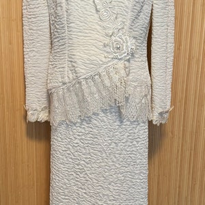 May include: A white dress suit with a lace trim and a floral design. The jacket has a V-neck and long sleeves. The skirt is knee-length and has a scalloped hem.