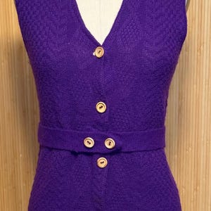 May include: A sleeveless, purple knit dress with a V-neck and a matching belt. The dress features a textured pattern and is fastened with wooden buttons. The dress is displayed on a mannequin.