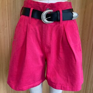 May include: Hot pink denim shorts with a black belt featuring a silver-colored buckle. The shorts have a high waist and pleats at the front. The belt has a decorative silver-colored buckle and a matching end piece.