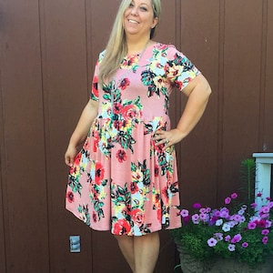 May include: A woman wearing a pink floral dress with short sleeves and a round neckline. She is standing on a wooden deck with a flower pot in the background.