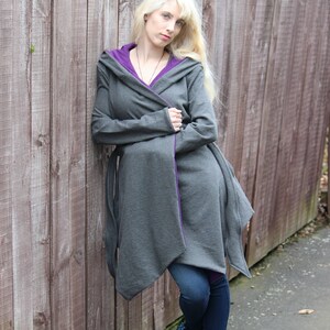 May include: A gray hooded cardigan with purple trim, worn by a person with long blonde hair. The cardigan is open and the person is leaning against a wooden fence.