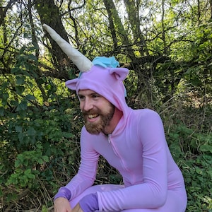 Könnte beinhalten: Eine Person trägt einen rosafarbenen Einhorn-Overall mit einem weißen Horn und einer blauen Mähne. Die Person hockt in einem grasbewachsenen Bereich.