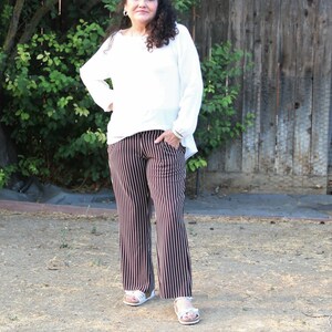 May include: A woman wearing a white long-sleeved shirt and striped pants. She is standing in front of a wooden fence.