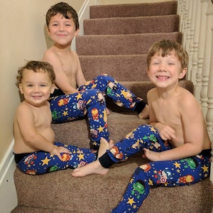 May include: Three children wearing blue pajama pants with a pattern of stars and superhero characters. The pajama pants have black cuffs. The children are sitting on a brown carpeted staircase.