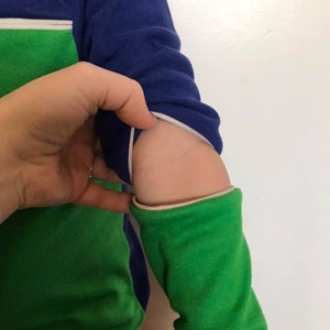 K&ouml;nnte beinhalten: Ein lang&auml;rmliges Shirt in Gr&uuml;n und Blau mit einem wei&szlig;en Rei&szlig;verschlussdetail am &Auml;rmel. Der Rei&szlig;verschluss ist heruntergezogen, wodurch der gr&uuml;ne Stoff darunter sichtbar wird.