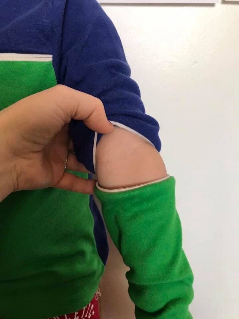 K&ouml;nnte beinhalten: Ein lang&auml;rmliges Shirt in Gr&uuml;n und Blau mit einem wei&szlig;en Rei&szlig;verschlussdetail am &Auml;rmel. Der Rei&szlig;verschluss ist heruntergezogen, wodurch der gr&uuml;ne Stoff darunter sichtbar wird.