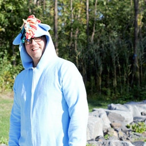 Può includere: Una persona indossa una tuta blu con cappuccio e un corno di unicorno colorato sul cappuccio. La persona sorride e guarda la telecamera. Lo sfondo è un'immagine sfocata di alberi e rocce.