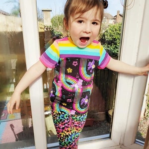 K&ouml;nnte beinhalten: Ein kleines Kind tr&auml;gt ein schwarz-regenbogen gestreiftes Shirt mit einem Care Bears-Druck und regenbogenfarbene Leoparden-Leggings.