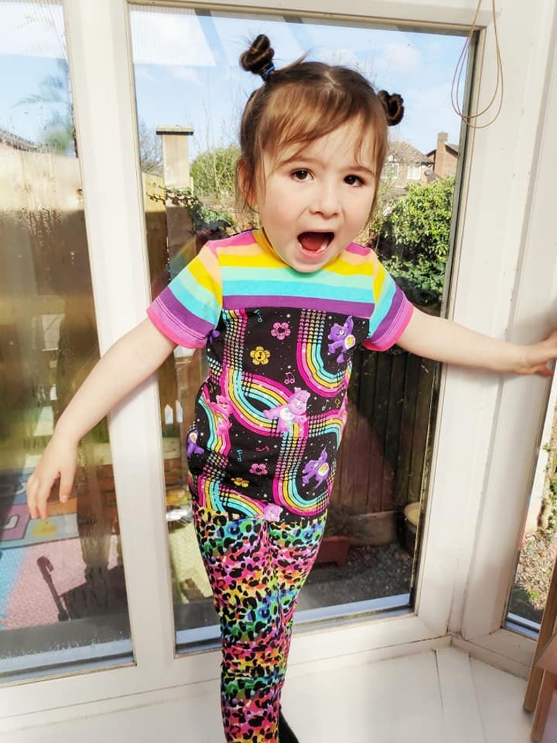 K&ouml;nnte beinhalten: Ein kleines Kind tr&auml;gt ein schwarz-regenbogen gestreiftes Shirt mit einem Care Bears-Druck und regenbogenfarbene Leoparden-Leggings.