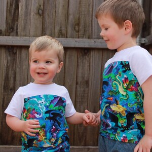 K&ouml;nnte beinhalten: Zwei junge Jungen tragen wei&szlig;e T-Shirts mit einem bunten Drachen-Druck auf der unteren H&auml;lfte. Die Jungen halten sich an den H&auml;nden und l&auml;cheln.