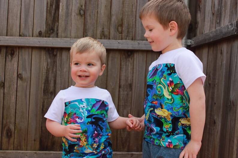 K&ouml;nnte beinhalten: Zwei junge Jungen tragen wei&szlig;e T-Shirts mit einem bunten Drachen-Druck auf der unteren H&auml;lfte. Die Jungen halten sich an den H&auml;nden und l&auml;cheln.