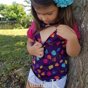 K&ouml;nnte beinhalten: Ein junges M&auml;dchen tr&auml;gt ein lilafarbenes und mehrfarbiges Shirt mit einem bunten Blatt-Muster, wei&szlig;e Shorts und eine blaue Blume im Haar. Das Shirt hat eine kleine &Ouml;ffnung vorne.
