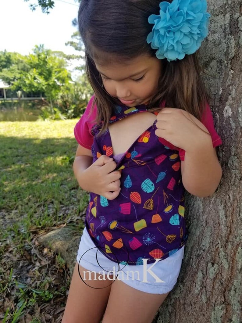 K&ouml;nnte beinhalten: Ein junges M&auml;dchen tr&auml;gt ein lilafarbenes und mehrfarbiges Shirt mit einem bunten Blatt-Muster, wei&szlig;e Shorts und eine blaue Blume im Haar. Das Shirt hat eine kleine &Ouml;ffnung vorne.
