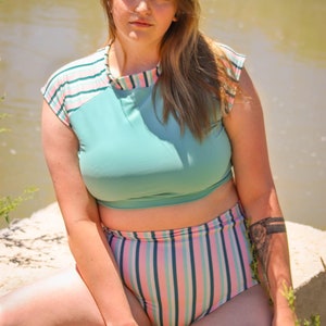 Puede incluir: Una mujer lleva un traje de baño de dos piezas a rayas turquesa y rosa. La parte superior es una camisa recortada de manga corta con un corpiño turquesa y mangas a rayas rosa y blancas. La parte inferior es una braga de bikini de talle alto a rayas rosa y blancas.