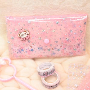 May include: A rectangular pink translucent pouch featuring a holographic star pattern and a panda-shaped pin on the front flap. Included are two rolls of patterned washi tape and a pink plastic wand with a bow and a circular frame.