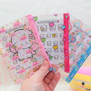Può includere: Tre bustine trasparenti in plastica con cerniera, decorate con stampe colorate di nuvole, cuori e arcobaleni. Ogni bustina riporta la scritta THECOFFEEMONSTERZCO e contiene un foglio di adesivi con illustrazioni e personaggi coordinati.