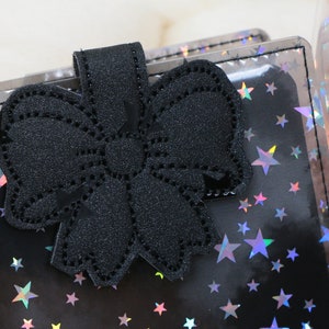 May include: A black notebook with a bow-shaped clasp. The notebook cover is black with a holographic star pattern. A clear tumbler with a cartoon face and glitter is in the background.