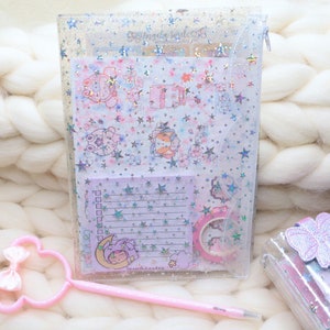 Può includere: Un raccoglitore di plastica trasparente con una finitura scintillante e una chiusura a zip. Il raccoglitore contiene diversi fogli di carta con un motivo a stelle e fiori. I fogli di carta hanno uno sfondo viola chiaro con una finitura olografica. Il raccoglitore è su una superficie bianca e soffice.