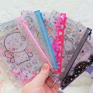 May include: A collection of five clear, glitter-filled vinyl pouches with colourful zippers in pink, blue, and black. Each pouch features a unique cartoon character illustration and loose sequins inside, creating a shaker effect for storing small items.