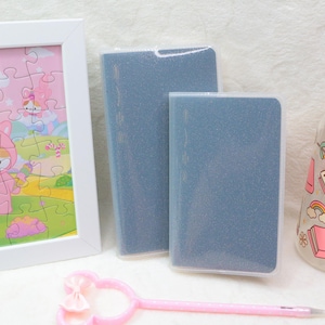 May include: A framed puzzle with a pink and yellow background and a cartoon unicorn character. Two blue notebooks, a glass jar with a wooden lid, and a pink pen are also in the image. The jar has a book and rainbow design.