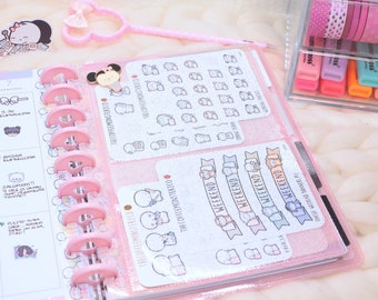May include: A pink disc-bound planner featuring a glittery cover, filled with various sticker sheets from The Coffee Monsterz Co. The planner includes pages with handwritten notes, cartoon character illustrations, and the text WEEKEND on banners.