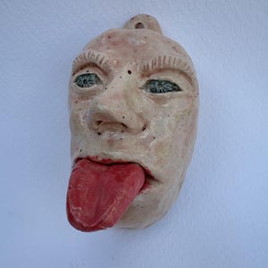 Può includere: Un'applique da parete in ceramica a forma di volto con la lingua di fuori. Il volto è rosa pallido con occhi blu e una lingua rossa. C'è un anello nella parte superiore per appenderlo. Scultura artigianale.
